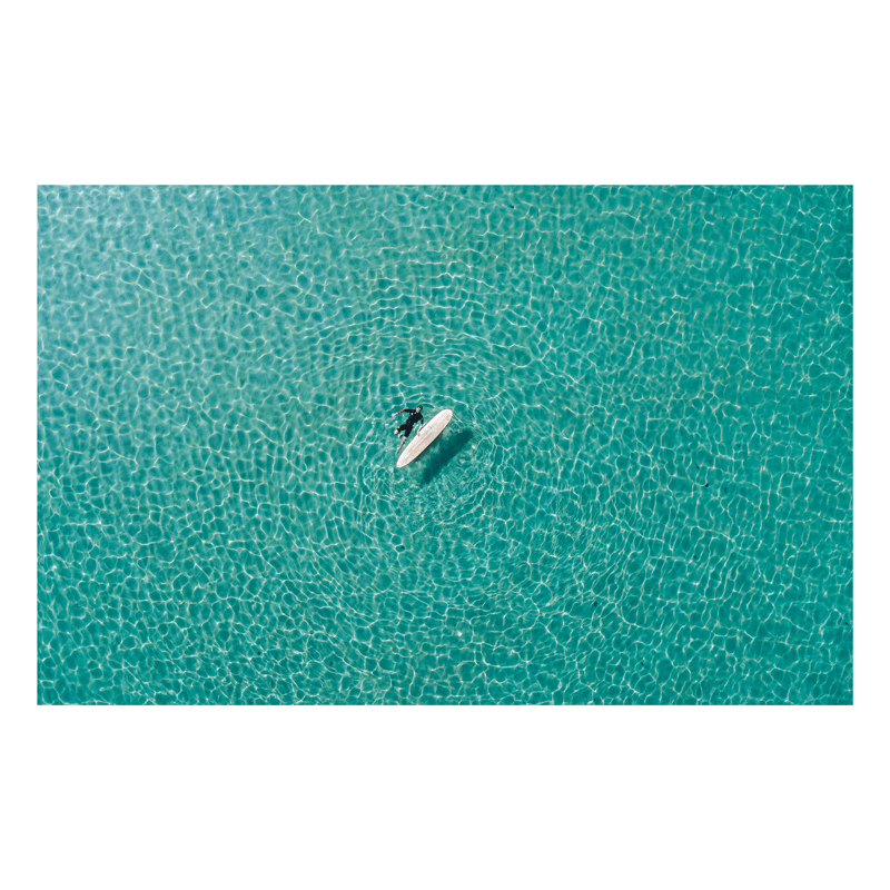 A breathtaking aerial view of a lone surfer floating in the crystal-clear waters of Forresters Beach, NSW, surrounded by sunlit ripples.