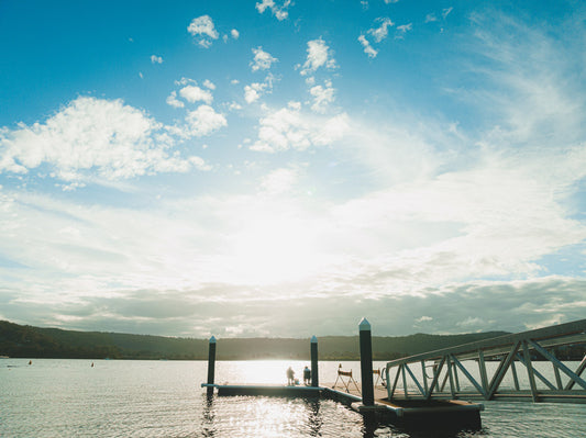 A tranquil scene at a jetty in Gosford, NSW, with two figures sitting by the water as the sun casts a golden reflection on the calm surface.
