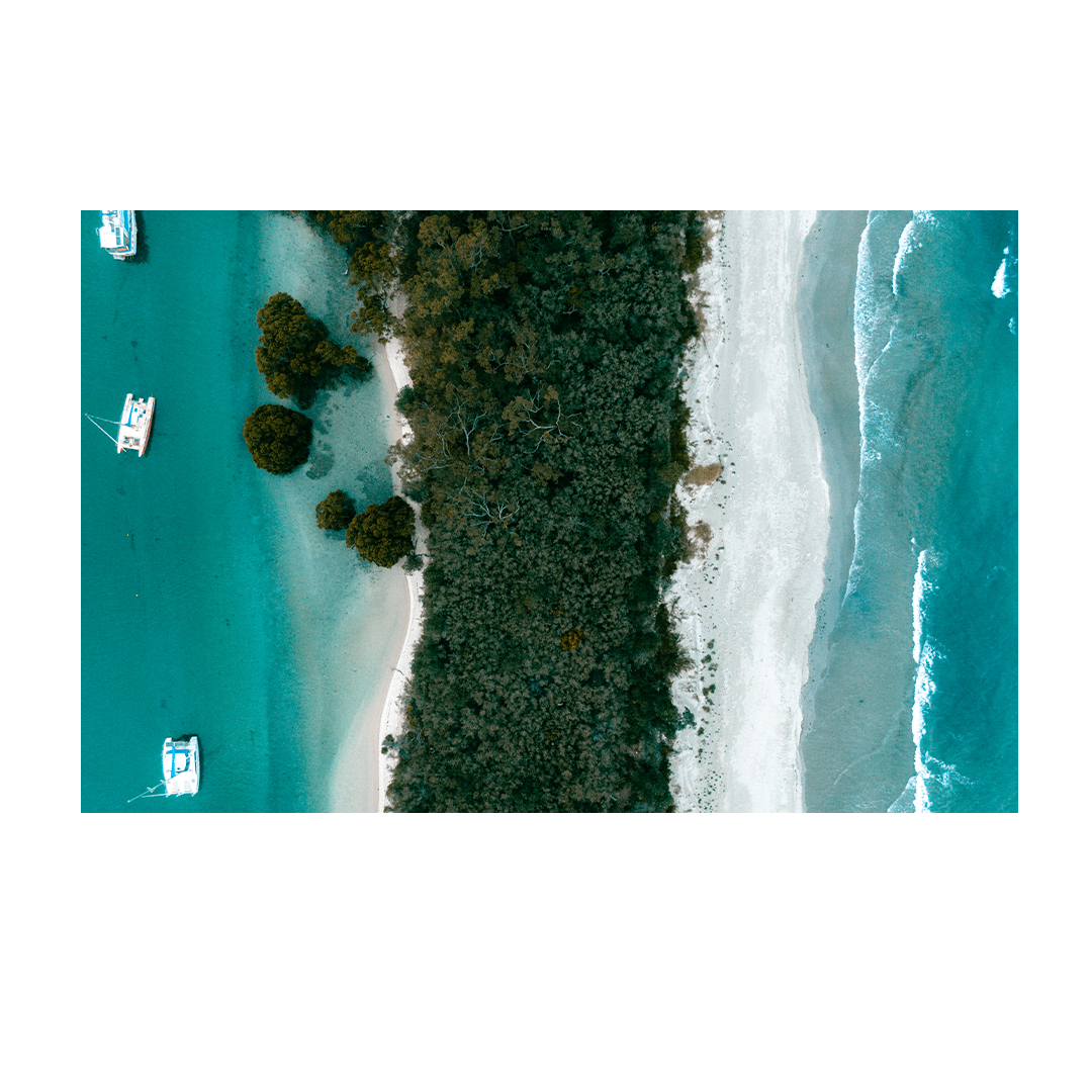 Aerial view of Huskisson on the South Coast of NSW, showcasing a striking contrast between lush greenery, pristine white sand, and vibrant turquoise waters with anchored boats nearby.