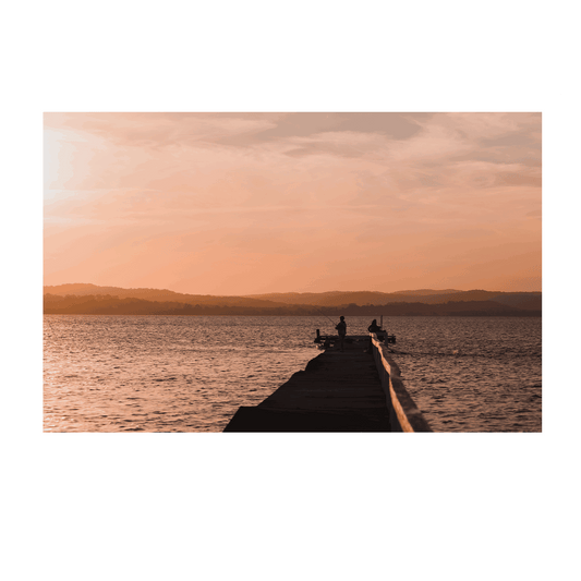 A tranquil sunset scene at Long Jetty, NSW, featuring silhouetted figures fishing against a golden sky reflecting on calm waters.
