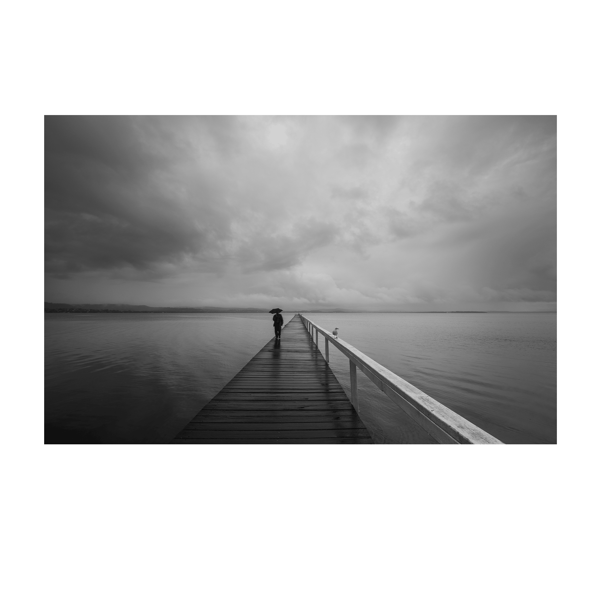 A striking black and white coastal scene of Long Jetty, NSW, featuring a lone figure walking with an umbrella along a rain-soaked boardwalk under stormy skies.