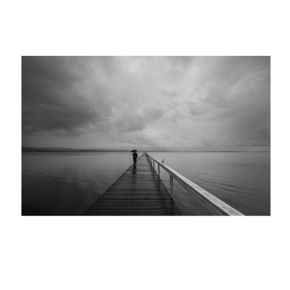 A striking black and white coastal scene of Long Jetty, NSW, featuring a lone figure walking with an umbrella along a rain-soaked boardwalk under stormy skies.