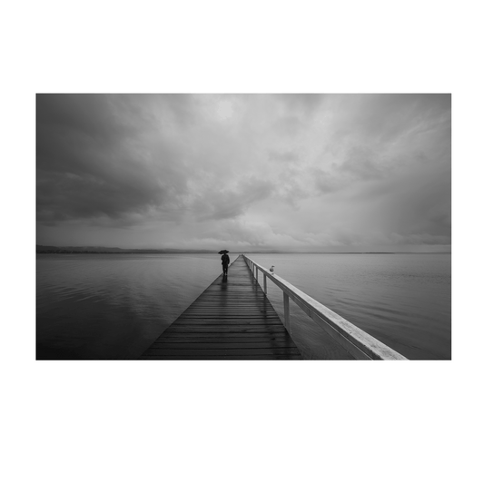 A striking black and white coastal scene of Long Jetty, NSW, featuring a lone figure walking with an umbrella along a rain-soaked boardwalk under stormy skies.