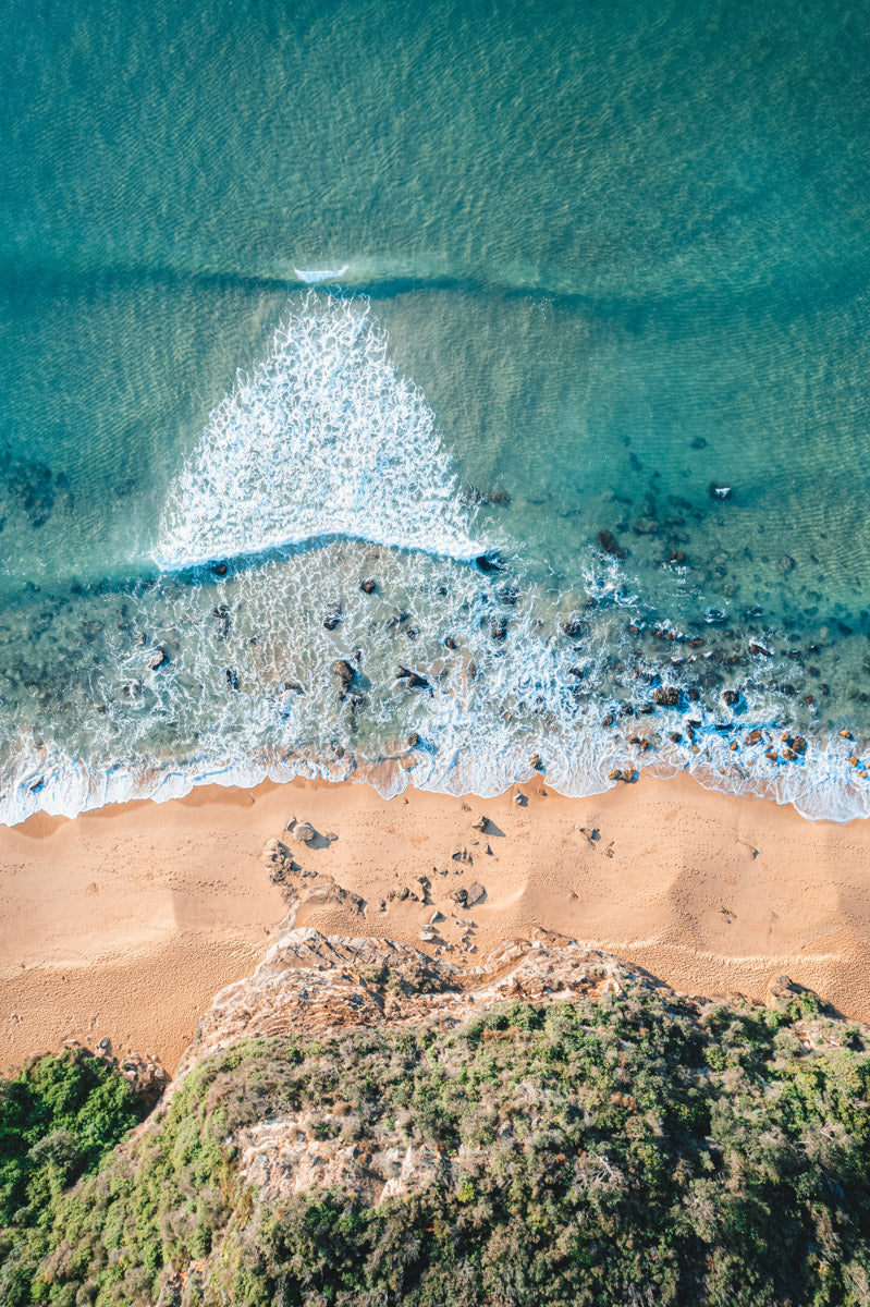 A stunning aerial view of Macmasters Beach, NSW, where waves converge in an arrow-like formation against golden sands and rocky shoreline.