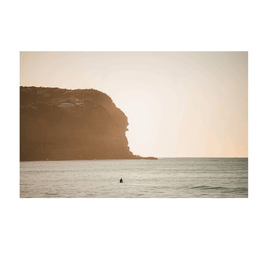 A lone surfer waits in the golden sunrise at Macmasters Beach, NSW, with a towering cliff bathed in soft morning light.
