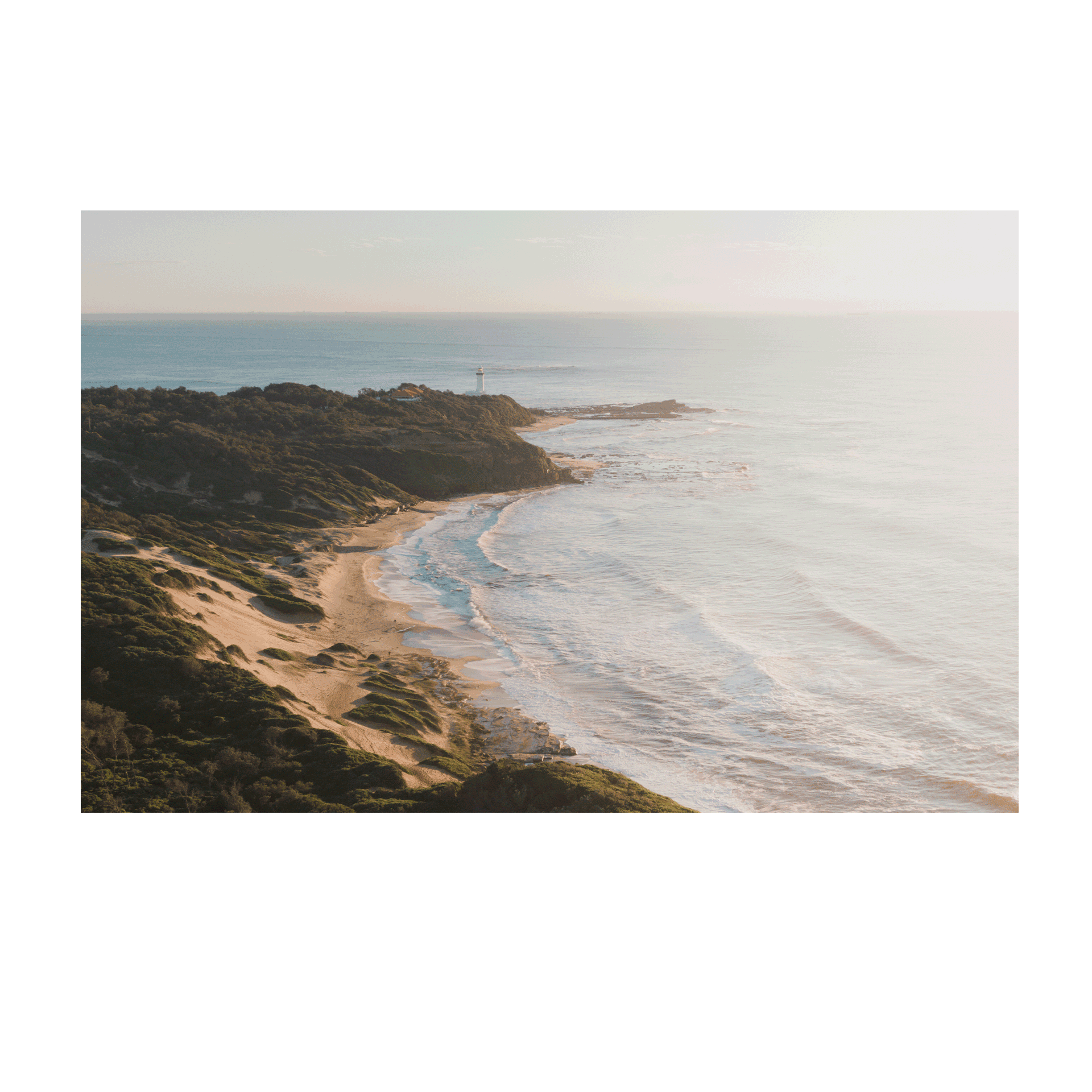 A soft, misty aerial view of Norah Head, NSW, featuring rolling sand dunes, a rugged coastline, and gentle ocean waves bathed in morning light.