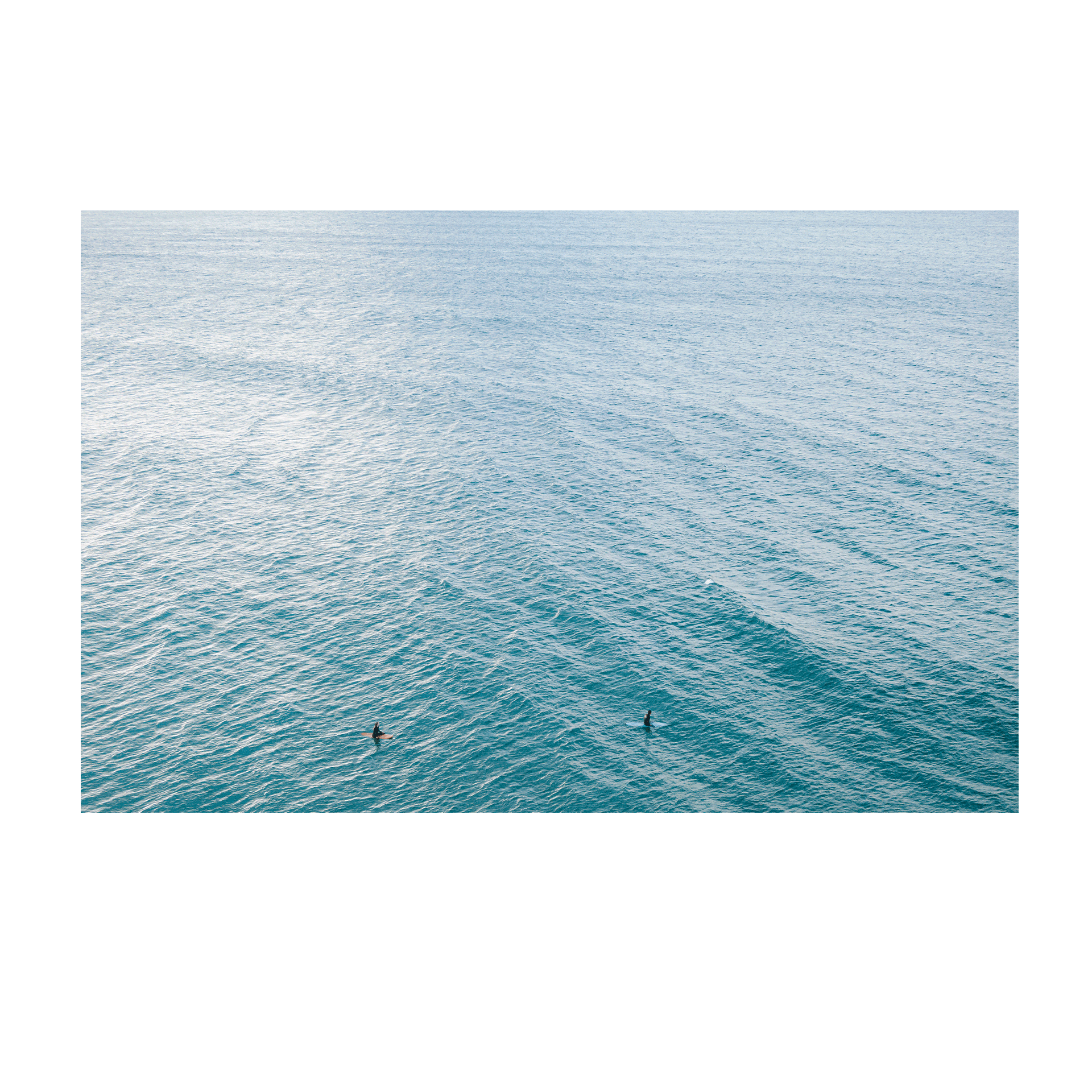 A peaceful aerial view of Norah Head, NSW, featuring two surfers floating in the vast, rippling blue ocean.
