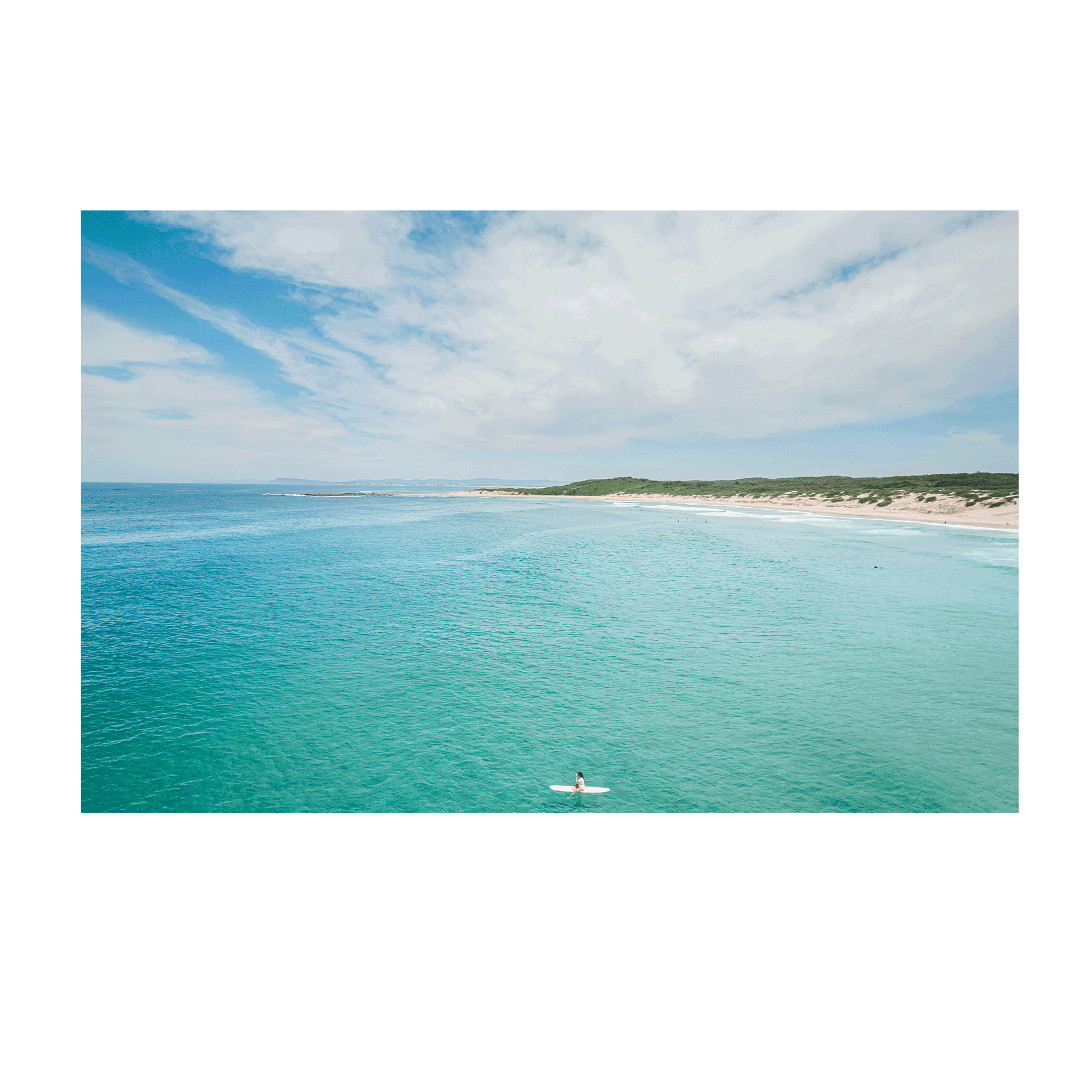 A breathtaking coastal aerial view of Soldiers Beach, Norah Head, NSW, featuring a lone surfer in turquoise waters with rolling dunes in the background.
