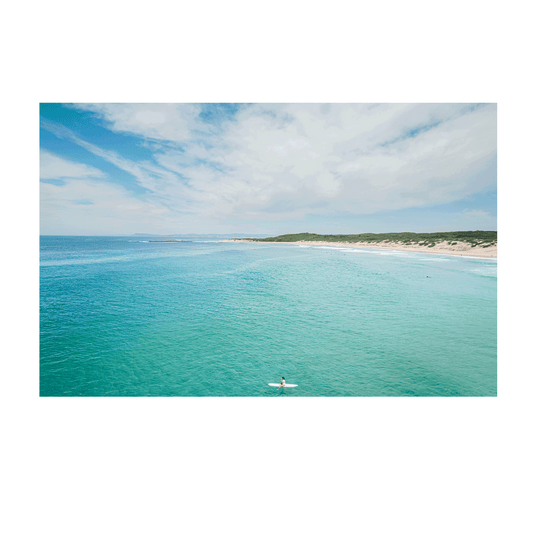 A breathtaking coastal aerial view of Soldiers Beach, Norah Head, NSW, featuring a lone surfer in turquoise waters with rolling dunes in the background.
