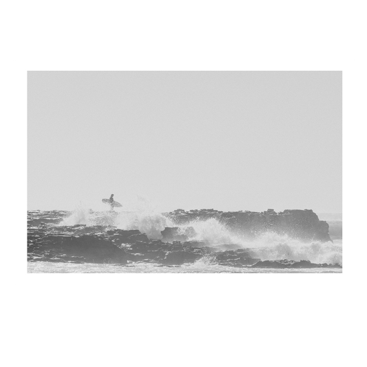 Black and white coastal print of a surfer navigating the rugged shoreline of North Avoca Beach, NSW, with waves crashing against the rocky outcrop.