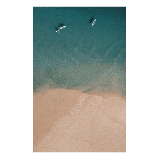 An aerial view of Red Rock, NSW, showcasing turquoise waters and sandy beaches, with two paddleboarders gliding across the bay.
