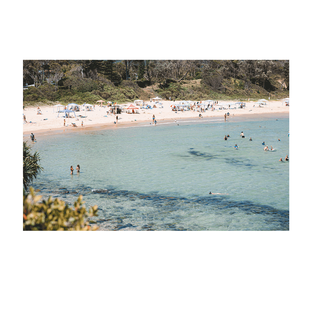 A lively summer scene at Scotts Head, NSW, featuring clear turquoise waters, swimmers enjoying the refreshing ocean, and sunbathers relaxing along the golden sandy beach. The vibrant coastal setting captures the essence of a perfect beach day.
