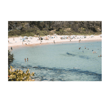 A lively summer scene at Scotts Head, NSW, featuring clear turquoise waters, swimmers enjoying the refreshing ocean, and sunbathers relaxing along the golden sandy beach. The vibrant coastal setting captures the essence of a perfect beach day.
