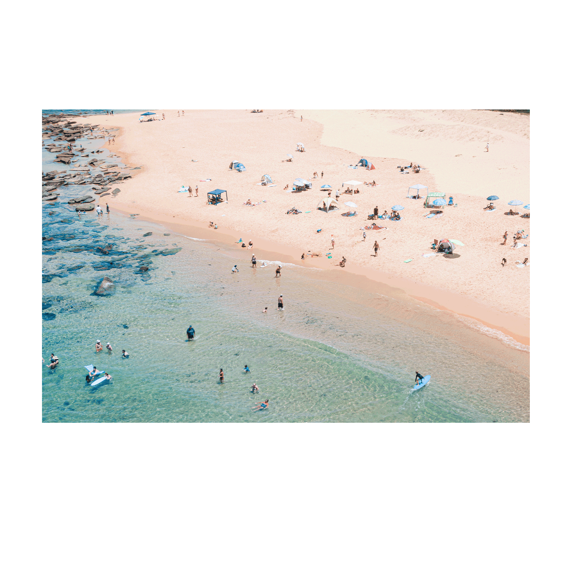 An aerial view of Spoon Bay, Central Coast, filled with beachgoers enjoying golden sands, scattered umbrellas, and turquoise waters on a perfect summer day.