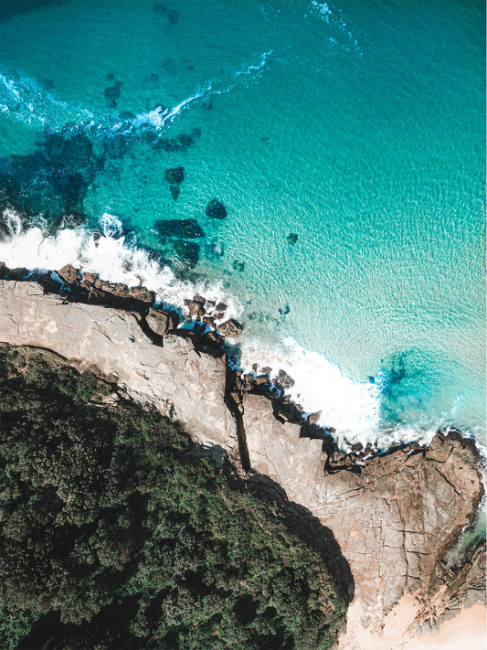 An aerial view of Spoon Bay, Central Coast, showcasing deep turquoise waters crashing against rugged rock formations with lush coastal greenery.
