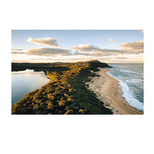 An aerial view of Spoon Bay, Central Coast, bathed in golden light, featuring rolling dunes, lush headlands, and deep blue ocean waves under a soft sunset sky.