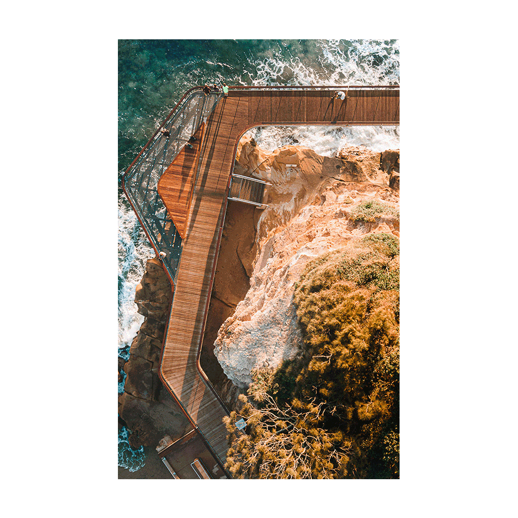 Aerial view of the Terrigal boardwalk winding along the coastal cliffs, with waves crashing below and golden sunlight highlighting the rugged landscape.
