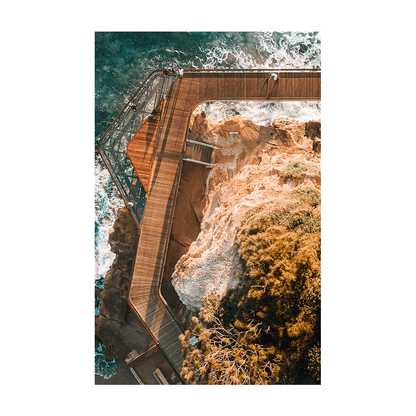 Aerial view of the Terrigal boardwalk winding along the coastal cliffs, with waves crashing below and golden sunlight highlighting the rugged landscape.
