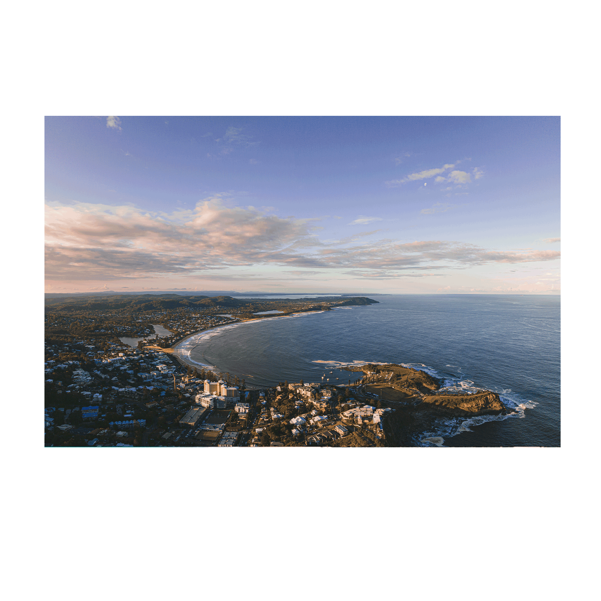 A breathtaking aerial view of Terrigal Beach, NSW, at golden hour, showcasing a vast ocean, rolling hills, and a pastel-hued sky with soft clouds.