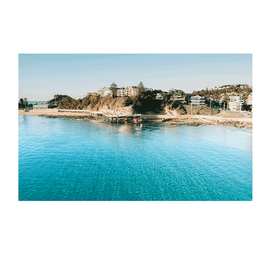 An aerial view of Terrigal Beach, NSW, showcasing crystal-clear turquoise waters, rugged cliffs, and beachfront homes under golden sunlight.
