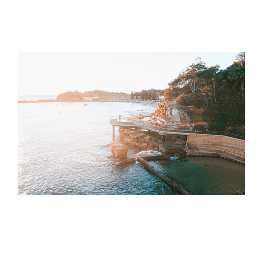 A sunlit coastal scene of the Terrigal boardwalk, NSW, glowing in warm golden light with gentle ocean waves and lush greenery in the background.