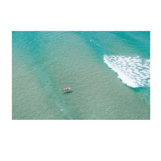 Aerial view of Toowoon Bay Beach, featuring a lone surfer floating on a board in crystal-clear turquoise waters. Gentle waves roll in, creating a peaceful and serene coastal scene. A perfect print for ocean lovers and beach enthusiasts.