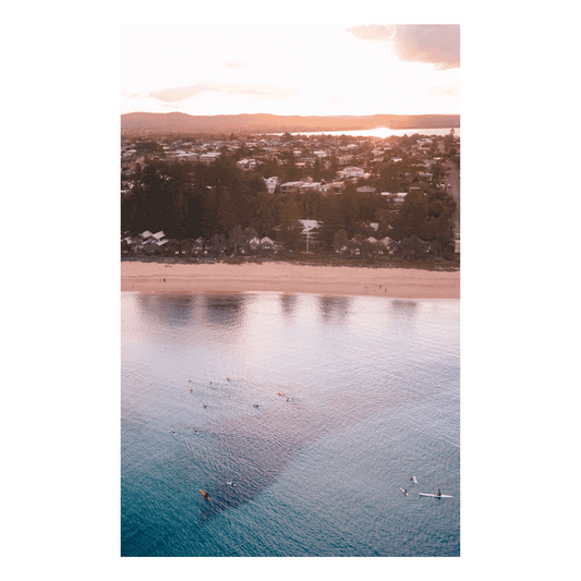 Aerial view of Toowoon Bay Beach at sunset, with golden light reflecting off calm waters. Paddleboarders and kayakers glide across the bay, creating a peaceful coastal scene perfect for a serene and modern space.
