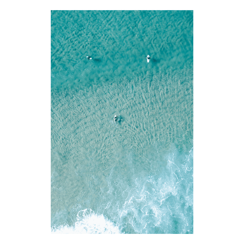 📸 An aerial view of Wamberal Beach, NSW, featuring three surfers in crystal-clear turquoise waters, with soft waves rolling onto the shore.
