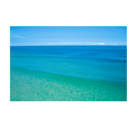 An aerial view of Wamberal Beach, NSW, featuring surfers floating in tranquil turquoise waters with a stunning gradient of deep blue ocean hues.
