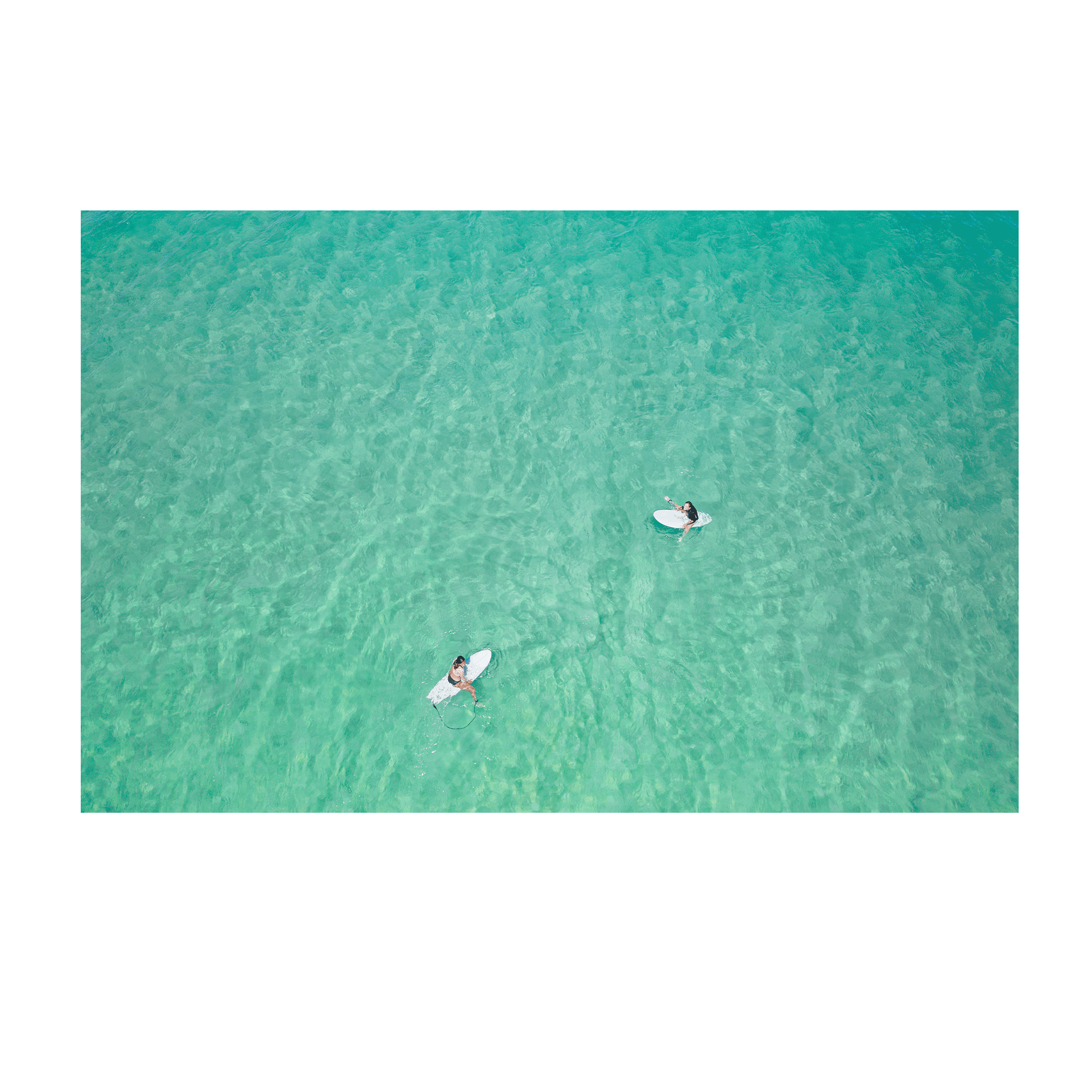 An aerial view of Wamberal Beach, NSW, showcasing two surfers floating in calm, crystal-clear turquoise waters.
