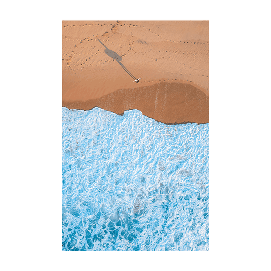 An aerial view of Wamberal Beach, NSW, featuring a lone surfer walking along golden sand, casting a long shadow as waves crash onto the shore.
